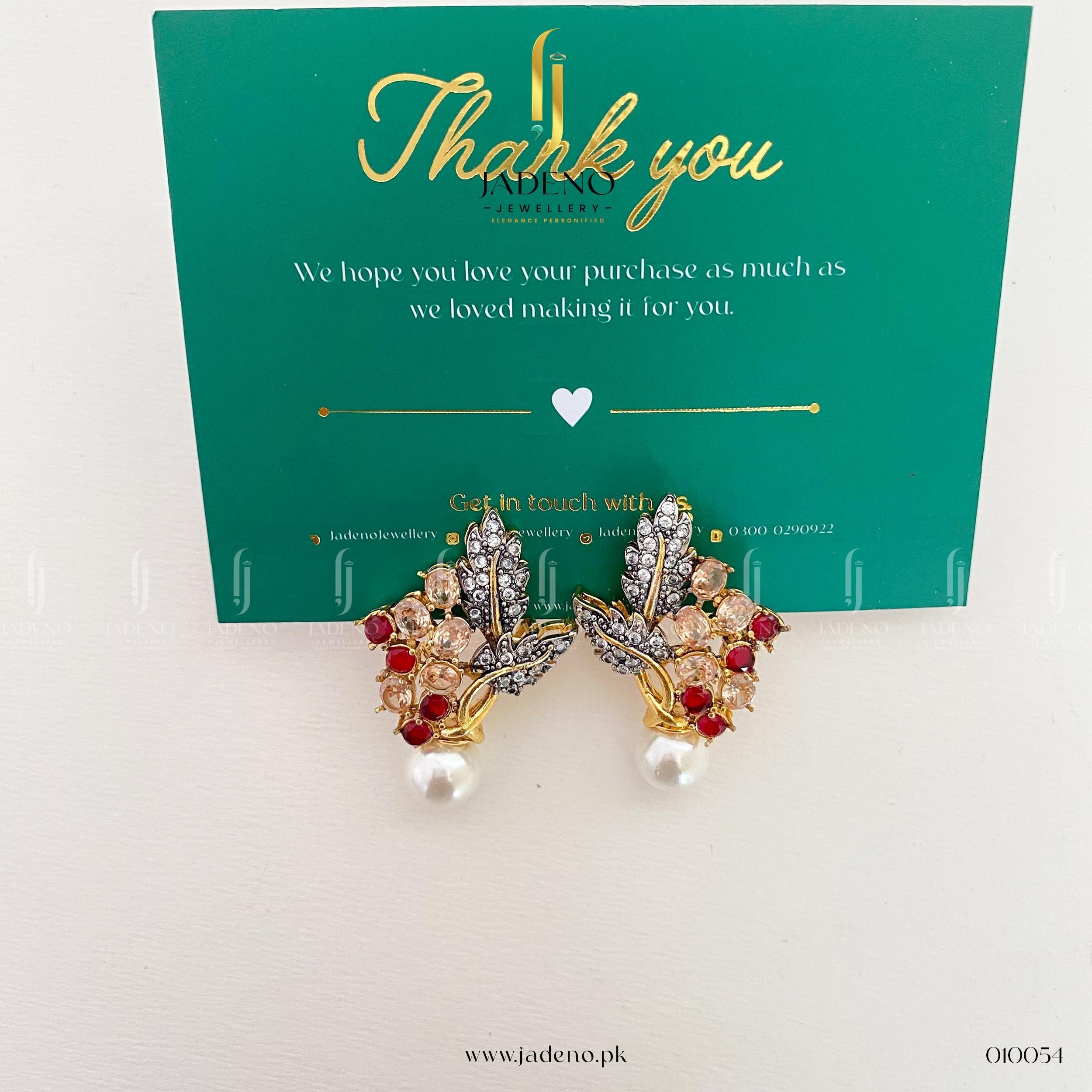 "Gold zircon stud earrings with red and clear stones and pearl drops displayed on a white surface with green background"
