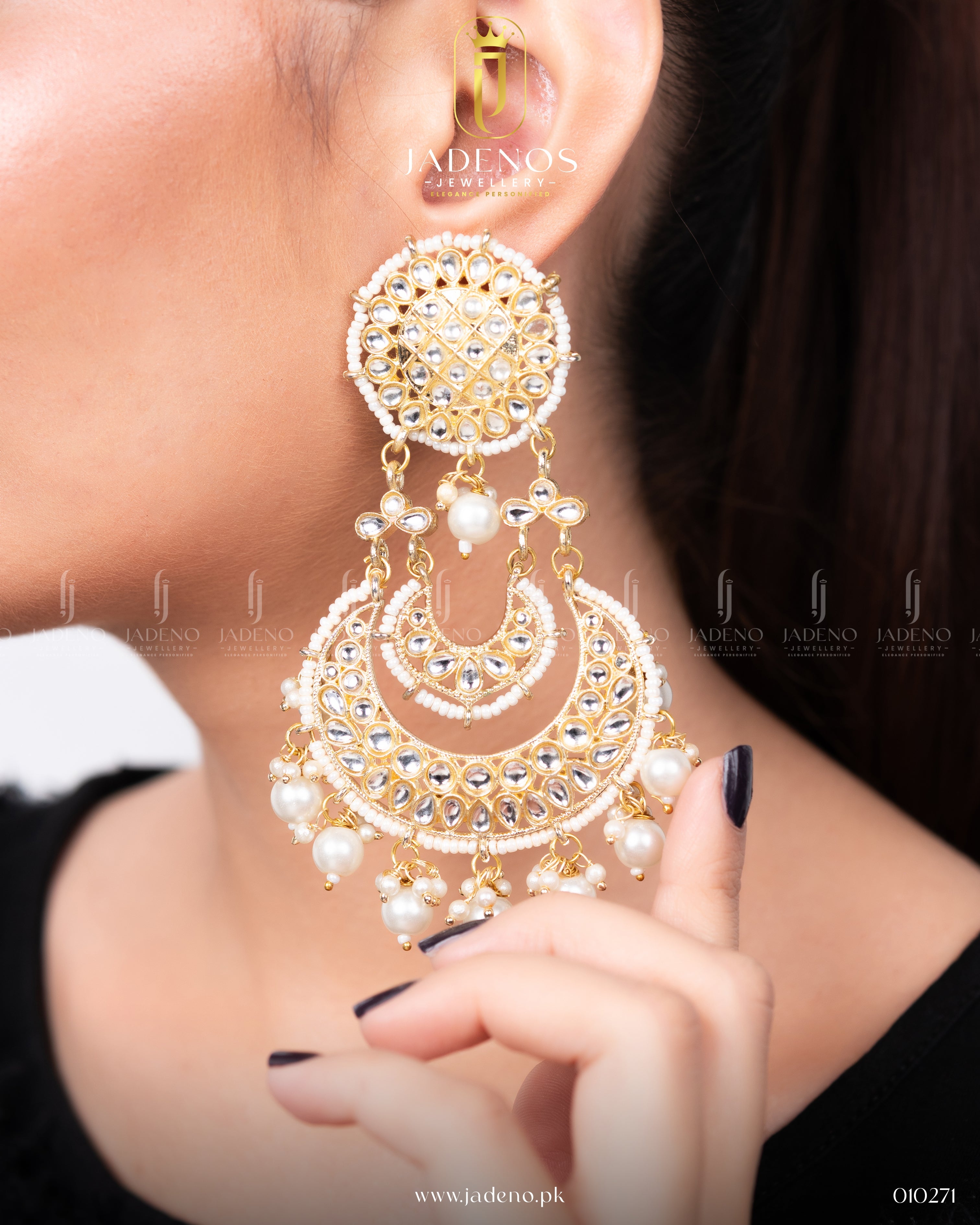 A pair of Zar-e-Noor Royal Chand Bali earrings featuring intricate designs with pearls and sparkling stones in a crescent moon shape, held by a hand with black nails.